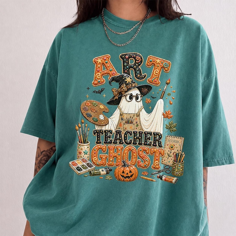 May include: A teal short-sleeve t-shirt featuring a graphic of a ghost wearing a witch hat and glasses, holding a paintbrush and palette. The text reads ART TEACHER GHOST, surrounded by art supplies, a pumpkin, and autumn leaves in orange and brown.