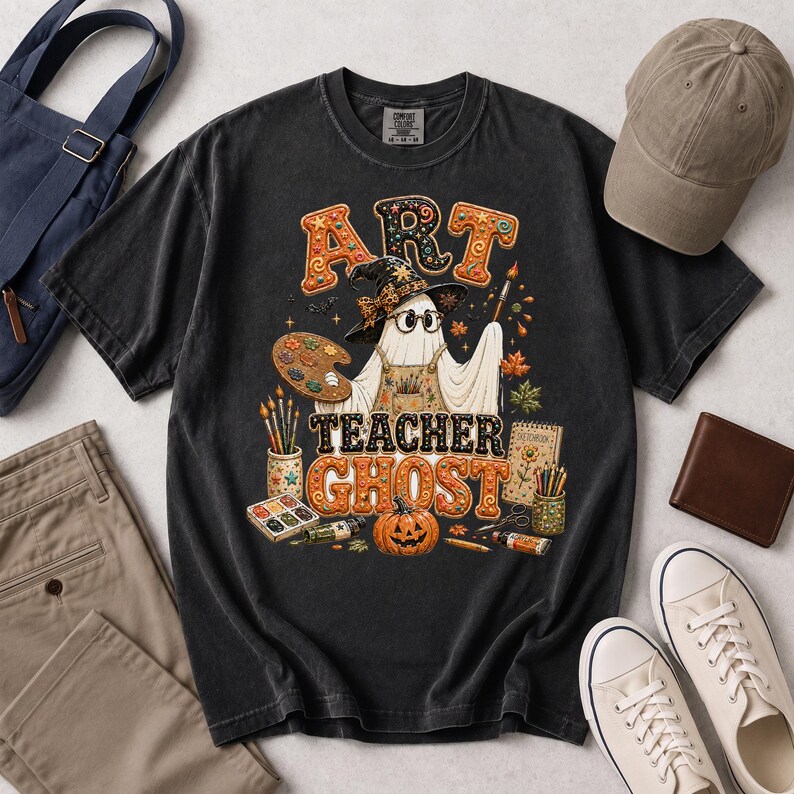 May include: A charcoal grey t-shirt featuring a graphic of a ghost wearing a witch hat and glasses, holding a paintbrush and palette. The text reads ART TEACHER GHOST, surrounded by art supplies, autumn leaves, and a jack-o'-lantern pumpkin.