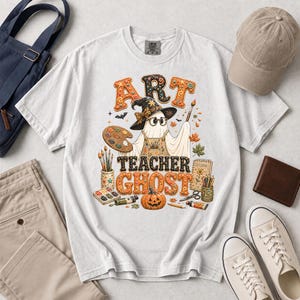 May include: A white t-shirt featuring a graphic of a ghost wearing a witch hat and glasses, holding a paintbrush. The text reads ART TEACHER GHOST, surrounded by art supplies, a pumpkin, and autumn leaves in orange, brown, and yellow tones.