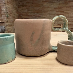 Pode incluir: Uma caneca grande de cerâmica rosa pálido com uma alça texturizada verde-clara, acompanhada por uma xícara pequena verde-clara salpicada e uma xícara menor rosa pálido lisa, todas com formas orgânicas e superfícies lisas.