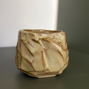Pode incluir: Uma pequena chávena de cerâmica com um exterior texturizado e facetado. A superfície apresenta um esmalte salpicado em verde claro e bege com detalhes em castanho escuro que realçam as cristas angulares irregulares e reentrâncias.