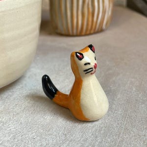 Pode incluir: Uma pequena figura de cerâmica de um gato com pêlo laranja e branco, orelhas pretas e cauda preta. O gato tem uma cara pintada com bigodes pretos e bochechas vermelhas.