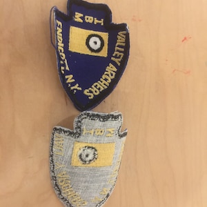 May include: Two embroidered patches in the shape of an arrowhead. The top patch is blue with yellow and white embroidery. The bottom patch is gray with yellow and white embroidery. The top patch reads "Valley Archers" and "Endicott, N.Y.". The bottom patch reads "Valley Archers".