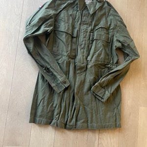 May include: A green military-style jacket with a button-up front and long sleeves. The jacket has two large pockets on the front and a small patch on the left sleeve with a red, white, and blue design.