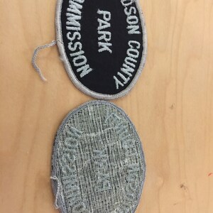 May include: Two oval-shaped embroidered patches. The top patch is black with white text that reads "HUDSON COUNTY PARK COMMISSION". The bottom patch is gray with white text that reads "HUDSON COUNTY PARK COMMISSION".
