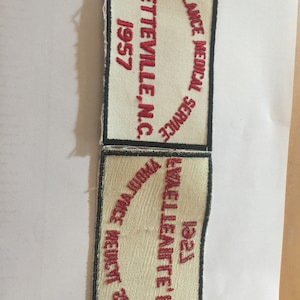 May include: Two white fabric patches with black borders. The top patch has red embroidered text that reads "AMBULANCE MEDICAL SERVICE FAYETTEVILLE, INC. 1957". The bottom patch has red embroidered text that reads "AMBULANCE MEDICAL SERVICE KNOXVILIE, TN, INC. 1927".