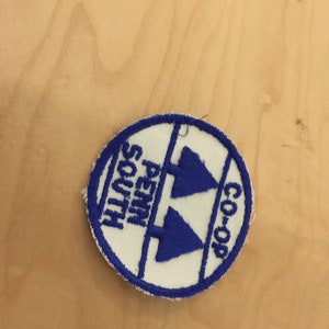 May include: A white circular patch with blue embroidered text and a blue embroidered design of two pine trees. The text reads "Penn South Co-op".