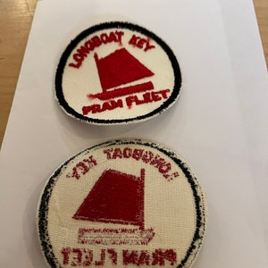 May include: Two round white fabric patches with black embroidered borders. The top patch has the text "Longboat Key Pram Fleet" and a red sailboat. The bottom patch has the text "Longboat Key Pram Fleet" and a red sailboat.