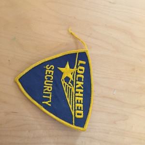 May include: A blue and yellow embroidered patch with the text "LOCKHEED SECURITY" and a star and wing design.