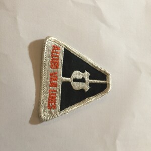 May include: A triangular black and white patch with orange text that reads "Rolling With Times". The patch features a white silhouette of a guitar with a single pick-up.