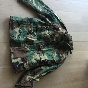 Puede incluir: Una chaqueta de camuflaje verde, marrón y negro con capucha con cordón y el texto "U.S. ARMY" en la manga izquierda.