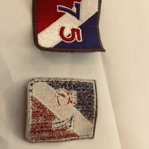 May include: Two embroidered patches with red, white, and blue colors. The top patch has the number "5" and a diagonal line with the number "7" on it. The bottom patch has a stylized design with a circle and a diagonal line.