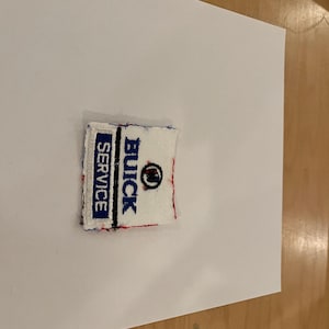 May include: A rectangular patch with the words "BUICK SERVICE" in blue and black lettering on a white background. The patch has a red border and a black Buick logo. The patch is likely for a Buick vehicle.