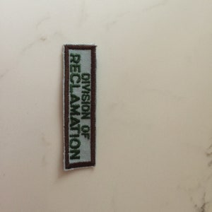 May include: A white patch with brown trim and green embroidered text that reads "Division of Reclamation".