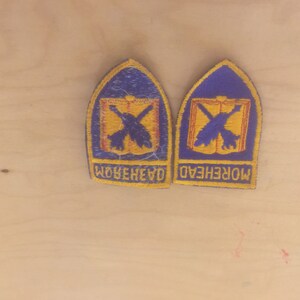May include: Two embroidered patches with a blue arched top and yellow border. Each patch features a yellow emblem with crossed arrows and a book. The word "MOREHEAD" is embroidered on the bottom.