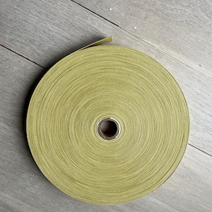 Peut inclure: Un rouleau enroulé de tissu vert olive clair, probablement un ruban ou un ruban adhésif, est posé sur une surface en bois gris clair. Le tissu est soigneusement enroulé, révélant un noyau en carton au centre. La texture du tissu est visible.