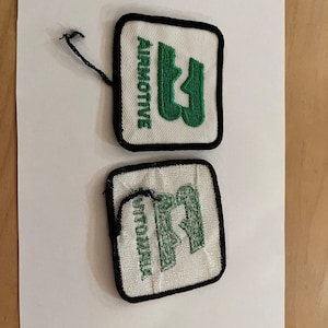 May include: Two square patches with black borders. The top patch has the word "AIRMOTIVE" in green, arched above a green stylized "U" shape on a white background. The bottom patch has the word "STOTUMIK" in green above a similar green "U" shape.