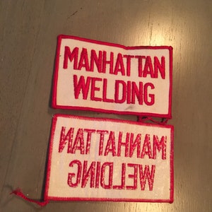 May include: Two white fabric patches with red embroidered text. The top patch reads "MANHATTAN WELDING". The bottom patch reads "WELDING MANHATTAN" in reverse.