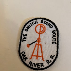 May include: White oval embroidered patch with black stitching. The text "The Switch Stand Route Oak River R.R." circles the edge. An orange stylized illustration of a railroad switch stand is in the center.