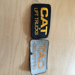 May include: Two embroidered patches, one black with yellow text reading "CAT LIFT TRUCKS" and one white with yellow text reading "TAC" with a smaller text reading "CONSTRUCTION".