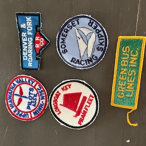 May include: Five embroidered fabric patches. One blue Denver & Roaring Fork RR Co patch, one circular Somerset Skipper Racing patch, one rectangular green Green Bus Lines Inc. patch, one circular Upper Hiawatha Valley Pilots Assn patch, and one circular Longboat Key Pram Fleet patch.