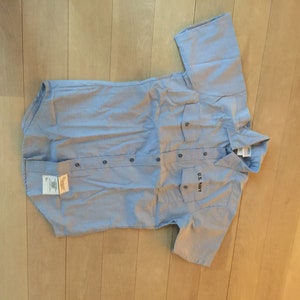 Puede incluir: Una camisa de manga corta azul claro con dos bolsillos y cierre de botones. La camisa tiene "U.S. NAVY" bordado en el bolsillo izquierdo.