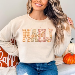 Peut inclure: Un sweat-shirt à col rond beige arborant le texte MAHJ en grandes lettres majuscules avec des motifs variés incluant du vichy orange, des taches de léopard, du tartan et des fleurs, avec le mot girl écrit en cursive au centre.
