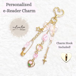 May include: A gold-colored e-reader charm with a heart-shaped clasp and a variety of dangling charms. The charms include pink beads, a star, a safety pin, and heart-shaped pendants. The text "Personalized e-Reader Charm" is visible.