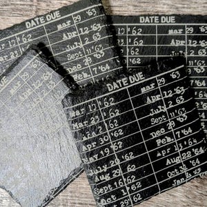 May include: A set of three square black slate coasters with rough, uneven edges. Each coaster features a white printed library book date due card design with columns for dates and years ranging from 1962 to 1965, including text reading DATE DUE.