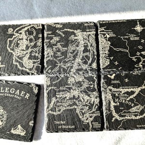May include: Set of six black slate coasters featuring maps of Middle-earth from The Lord of the Rings. Each coaster depicts a different region, including Gondor, Rohan, Mordor, and Belegaer (The Great Sea).