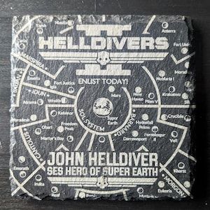 May include: A square, black slate coaster with white text and graphics. The coaster features the word "HELLDIVERS" above a skull, and a map of the Sol System. Text includes "ENLIST TODAY!" and "JOHN HELLDIVER SES HERO OF SUPER EARTH."