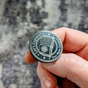 May include: A round, black pin with the words "Let's Explore!" and "One More Chapter!" encircling a central design. The design features an open book with a compass. The pin is held in a person's hand.