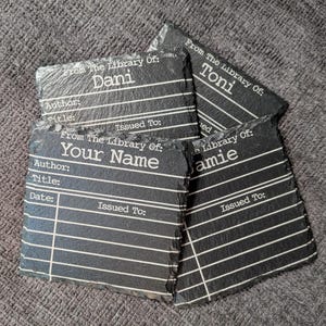 May include: A set of four square black slate coasters with rough, natural edges. Each coaster features a white printed library checkout card design with the text From The Library Of, Author, Title, Date, and Issued To, with names Dani, Toni, and amie.