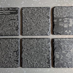 May include: A set of six square slate coasters featuring white laser-engraved text and maps. The designs include the Declaration of Independence, historical signatures, and a map of the thirteen colonies in America as of 1776 with text.