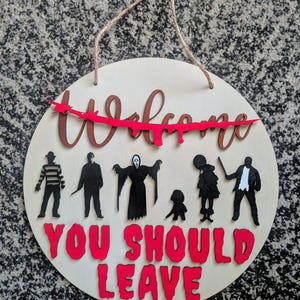 May include: Round wooden sign with the words "Welcome" and "You Should Leave" in red and brown lettering. The sign features silhouettes of horror movie characters and a red dripping accent. A jute rope is attached for hanging.