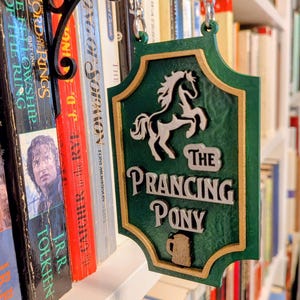 May include: A green, shield-shaped hanging sign with a gold border, featuring a raised white prancing horse silhouette, the text "THE PRANCING PONY" in white lettering, and a small brown mug icon at the bottom, suspended by a black metal bracket.