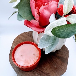 Op de afbeelding: Een rode kaars in een rode keramische houder met een roze was bovenkant. De kaars staat op een houten dienblad met een witte vaas met rode bloemen en groene bladeren.