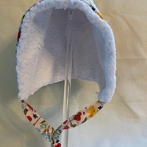 May include: A white, floral-patterned baby bonnet with a soft, white, fluffy interior. The bonnet features ties with a matching floral design. The exterior fabric displays red strawberries, yellow flowers, and blue accents on a cream background.