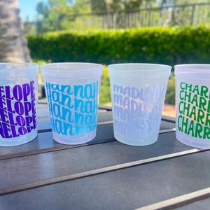 May include: Four translucent plastic cups arranged in a row. Each cup features a repeating name printed in a bold, stacked font: the first in purple with 'PENELOPE', the second in blue with 'hannah', the third in light blue with 'madison', and the fourth in green with 'CHARRETTE'.