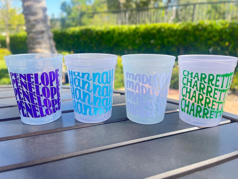 May include: Four translucent plastic cups arranged in a row. Each cup features a repeating name printed in a bold, stacked font: the first in purple with 'PENELOPE', the second in blue with 'hannah', the third in light blue with 'madison', and the fourth in green with 'CHARRETTE'.