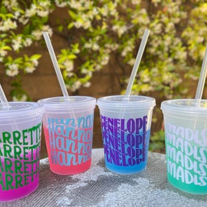 May include: Four clear plastic cold cups with lids and straws, each featuring a repeating name in a bold, wavy font. The cups contain colored liquids: pink with green text reading CHARRETTE, pink with blue text reading hannah, blue with purple text reading PENELOPE, and green with white text reading madison.