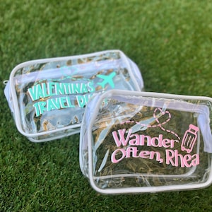 May include: Two clear travel pouches with white trim, sitting on green grass. One pouch has the text "Valentina's Travel Duo" in teal, with a teal airplane graphic. The other pouch has the text "Wander Often, Rhea" in pink, with a pink suitcase graphic.