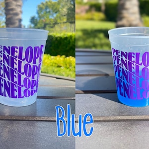 May include: A frosted plastic cup featuring the name Penelope printed in a repeating, wavy purple font. The cup is shown empty on the left and filled with a bright blue liquid on the right, demonstrating a color-changing effect when cold.