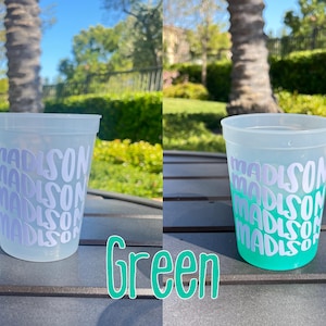 May include: A translucent plastic stadium cup featuring the name Madison repeated in white lettering. The cup on the left is clear, while the cup on the right shows a color-changing effect, transitioning to a vibrant green hue at the base.