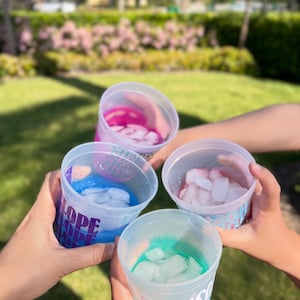 May include: Four translucent plastic cups held together in a circle, each containing a different colored liquid and ice cubes. The cups feature printed text in various fonts and colors, including the words SHORE, HOPE, and MADISON.