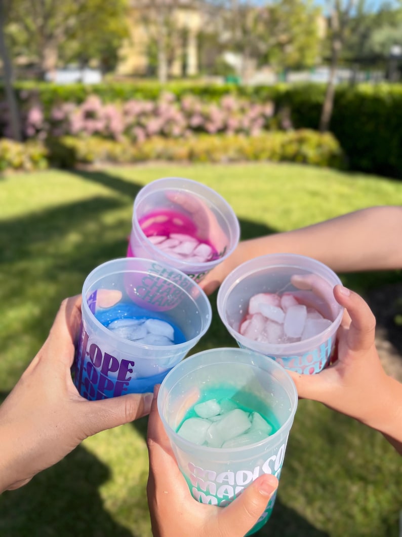 May include: Four translucent plastic cups held together in a circle, each containing a different colored liquid and ice cubes. The cups feature printed text in various fonts and colors, including the words SHORE, HOPE, and MADISON.