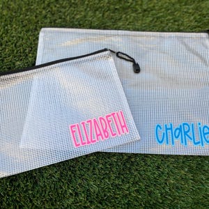 May include: Two clear mesh zipper pouches, one large and one small, with black zippers and pull cords. The smaller pouch has the name "ELIZABETH" in pink, and the larger pouch has the name "CHARLIE" in blue. The pouches are on a green surface.