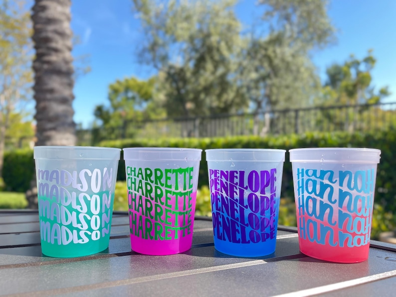 May include: Four translucent plastic cups with repeating names printed in bold, wavy typography. The first cup is teal with white text, the second is hot pink with green text, the third is blue with purple text, and the fourth is coral with blue text.