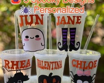 May include: Five clear plastic cups with lids and straws, each personalized with a name in orange lettering and a spooky Halloween design. Designs include a ghost, witch boots, bat, jack-o'-lantern, and spider.