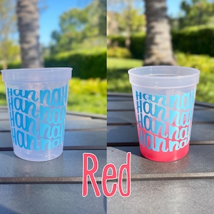 May include: A clear plastic cup featuring the name Hannah repeated in a repeating teal blue font pattern. The cup is shown empty on the left and filled with a red liquid on the right, with the word Red written in pink below the cups.